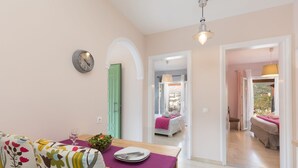 A dining area with a table set for two, a clock on the wall, and a view into a bedroom with a bed and a window.