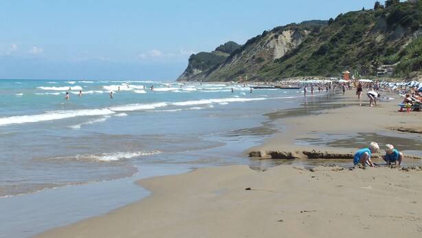 A beach with people swimming and walking along the shore, a sandy beach, and a rocky cliff in the background.