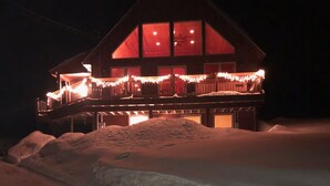 A snow-covered landscape with a wooden cabin featuring a balcony with string lights. - Cycladic Sunday Chalet (Bethel)