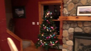A cozy living room with a wooden chair, a Christmas tree, a fireplace, and a stone wall. - Cycladic Sunday Chalet (Bethel)