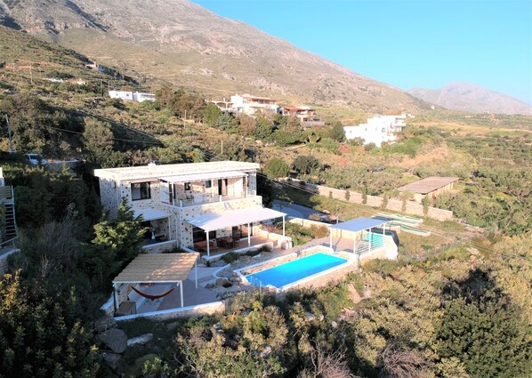 A two-story house with a pool, surrounded by greenery and mountains.