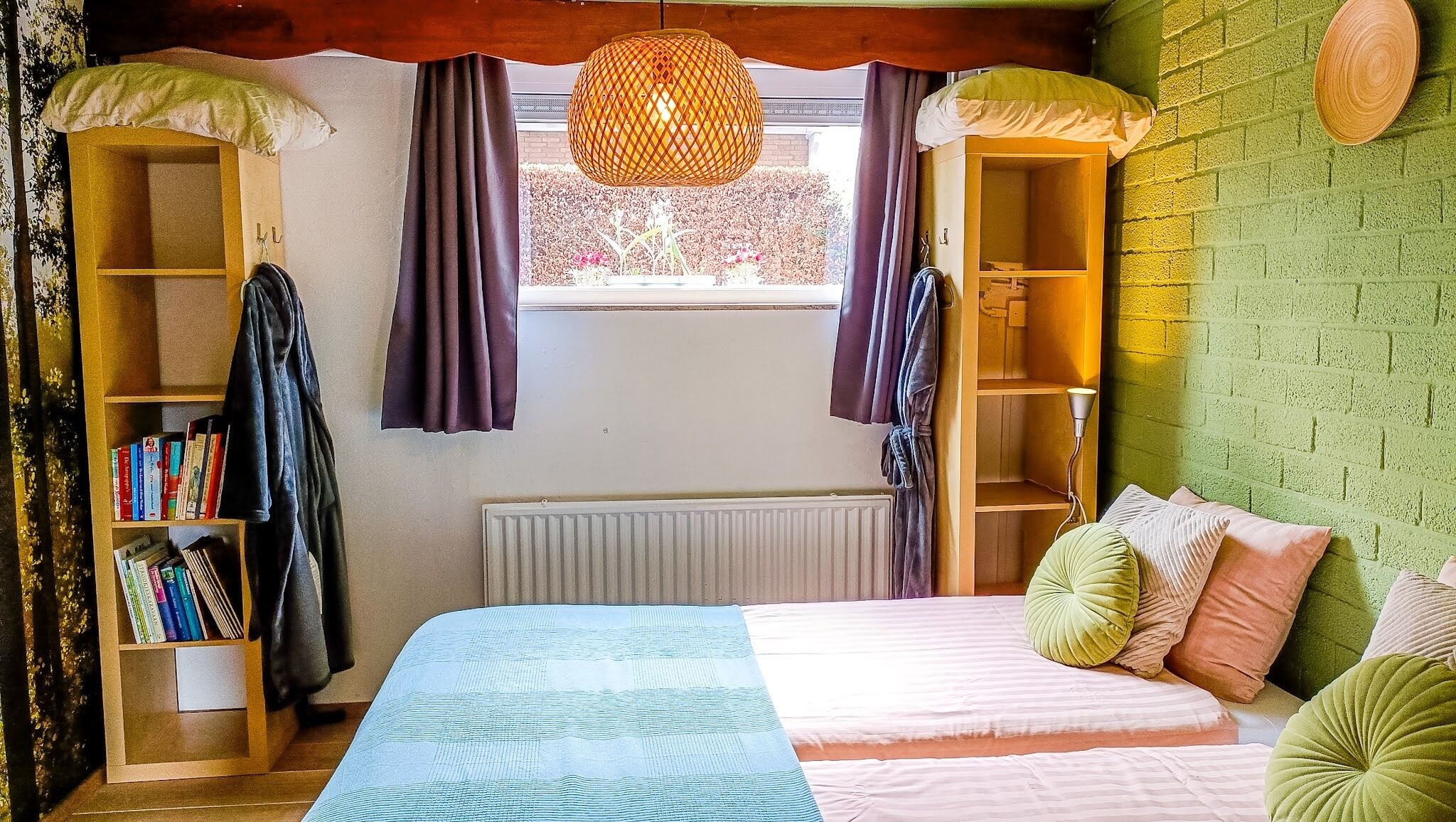 A bedroom with a bed, two bedside tables, a radiator, a window with curtains, and a green brick wall.
