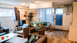 Ein modernes Wohnzimmer mit Essbereich, ausgestattet mit einer Couch, Sesseln, einem Tisch und einem großen Fenster mit Vorhängen.