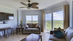 A living room with a sofa, a coffee table, a TV, and a ceiling fan. There are large windows with a view of greenery.