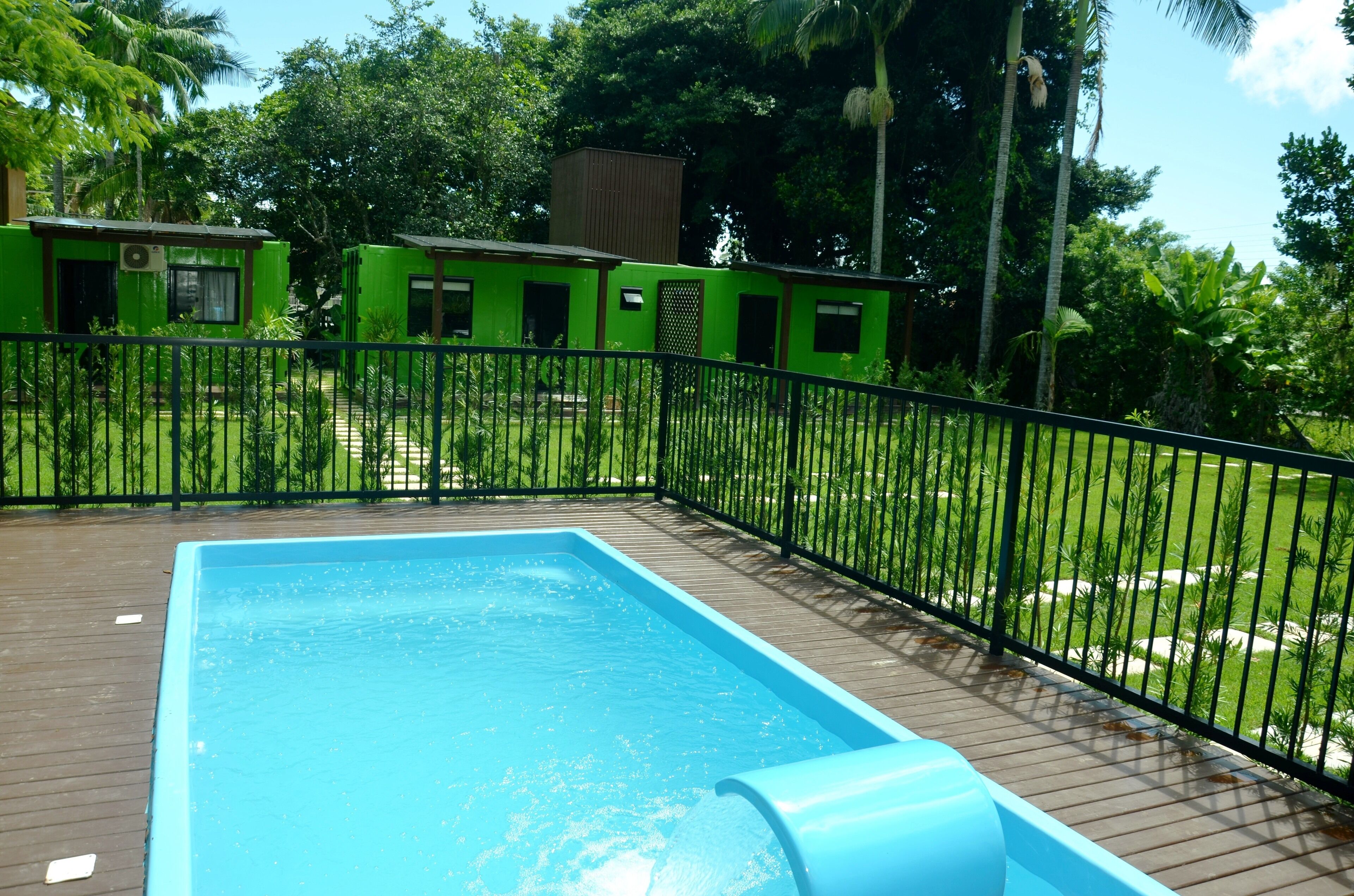 Ein Swimmingpool mit blauer Rutsche, umgeben von einer Holzterrasse und einem schwarzen Metallzaun.