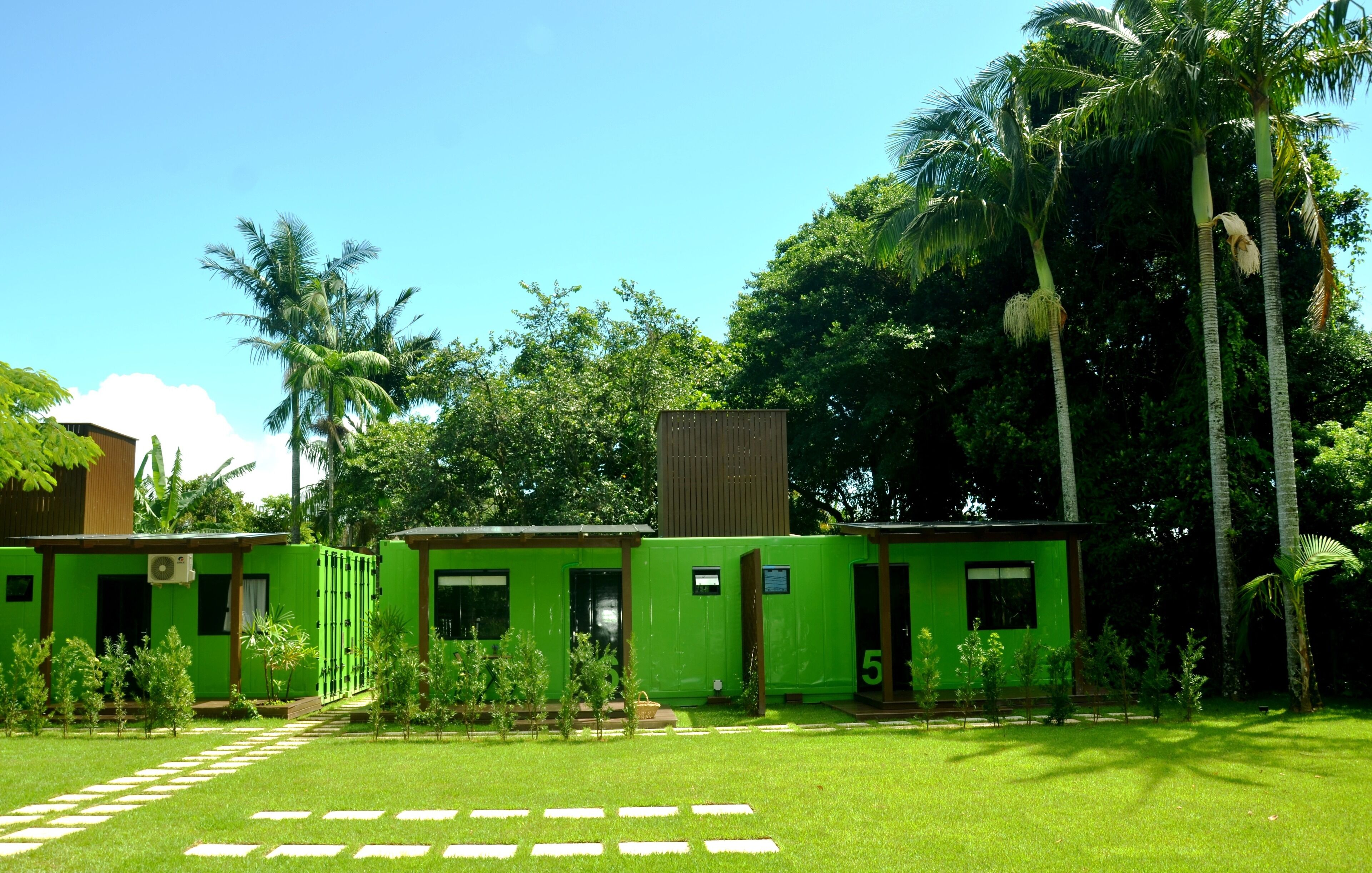 Una hilera de cabañas modulares verdes con un césped y palmeras al fondo.