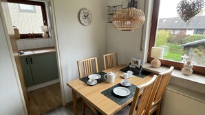Comedor con una mesa de madera dispuesta para cuatro personas, rodeada de sillas. Se ve una ventana con un adorno con forma de corazón, y una zona de cocina al fondo.