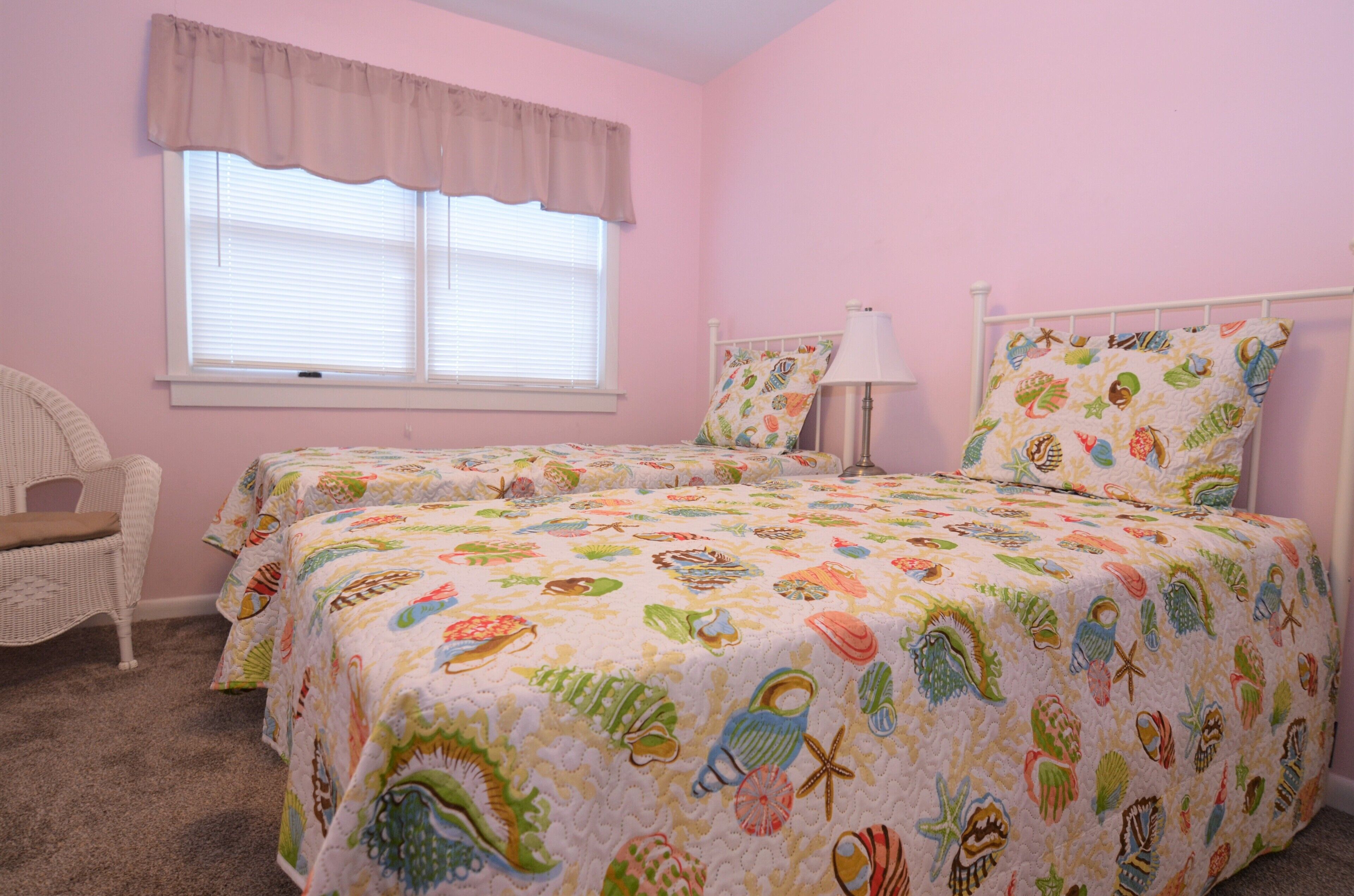 A pink bedroom with two beds, a chair, and a window with blinds.