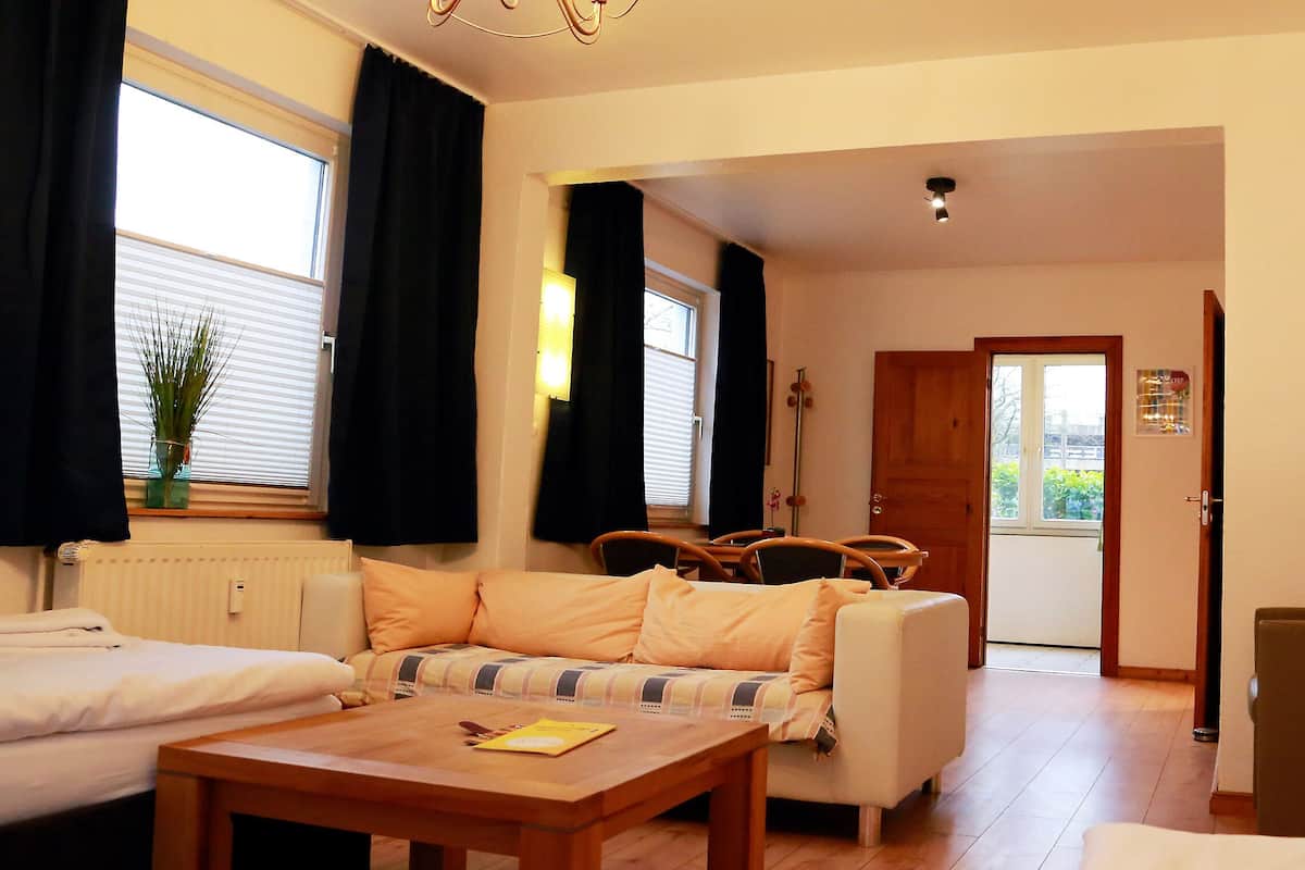 A cozy room with a sofa, a wooden table, and a chair. There is a window with curtains, a radiator, and a door leading to another room.