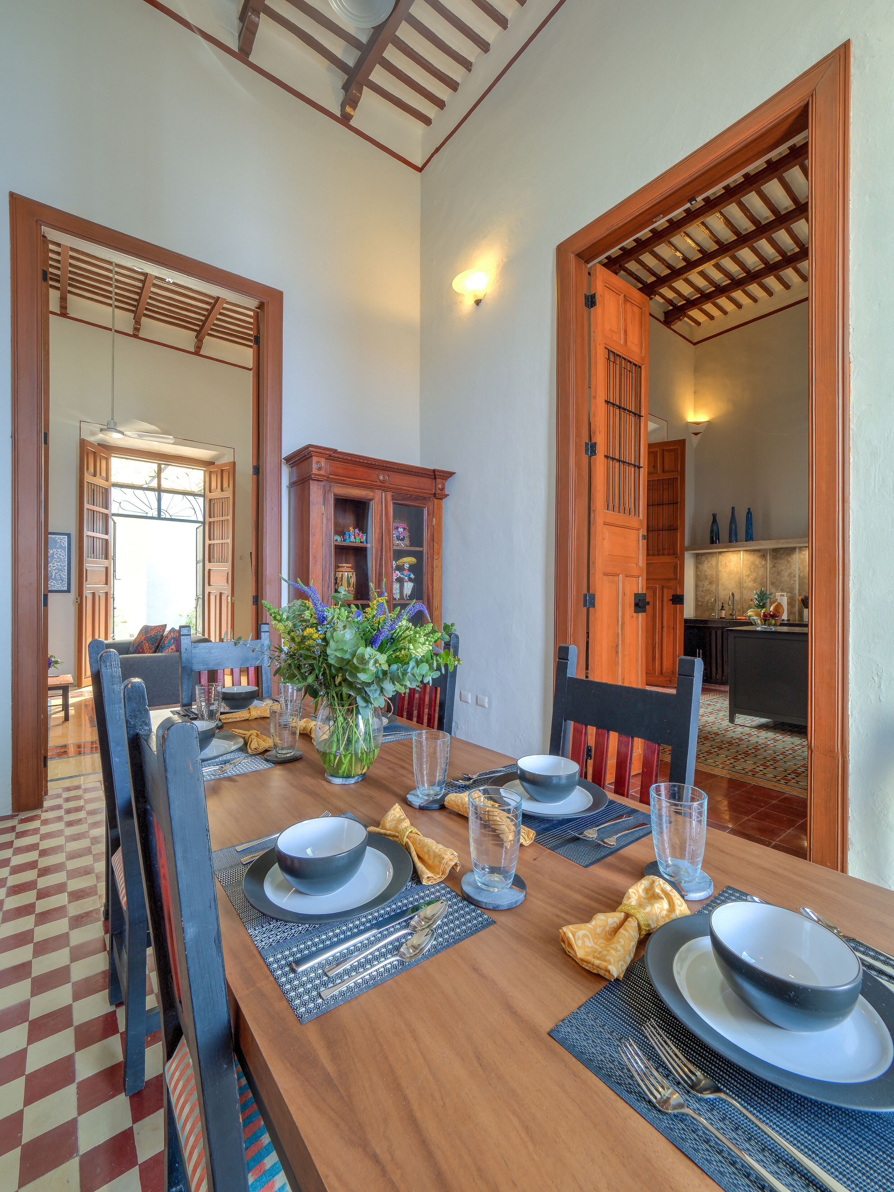 Un coin repas avec une table en bois, dressée pour quatre personnes, entourée de chaises. On aperçoit un meuble avec des portes en verre à l’arrière-plan.