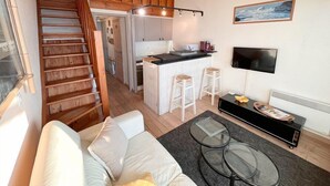 Um apartamento moderno com escada que leva a um mezanino, uma cozinha compacta, uma sala de estar com sofá e mesa de centro, e uma TV fixada na parede.