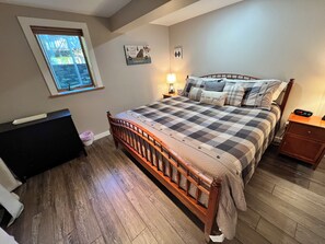 Una camera da letto con un letto in legno, un comodino, una cassettiera, una finestra vista alberi e un quadro appeso al muro.