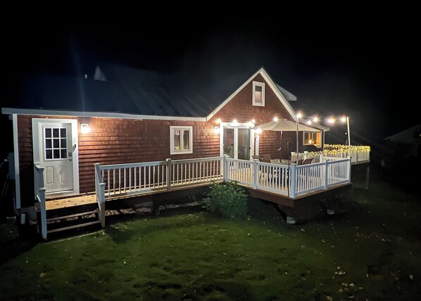Une maison en bois à deux étages, dotée d’un porche et d’une terrasse clôturée, éclairée par des lampes extérieures.
