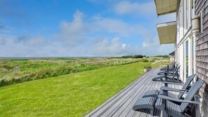 Une terrasse en bois, agrémentée de chaises, donnant sur une pelouse et un bâtiment doté d’un toit en pente.