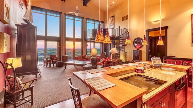 Un vaste espace de vie comprenant un grand salon avec une table à manger en bois, une cuisine dotée d’un îlot central et une vue sur l’extérieur grâce à de grandes fenêtres.
