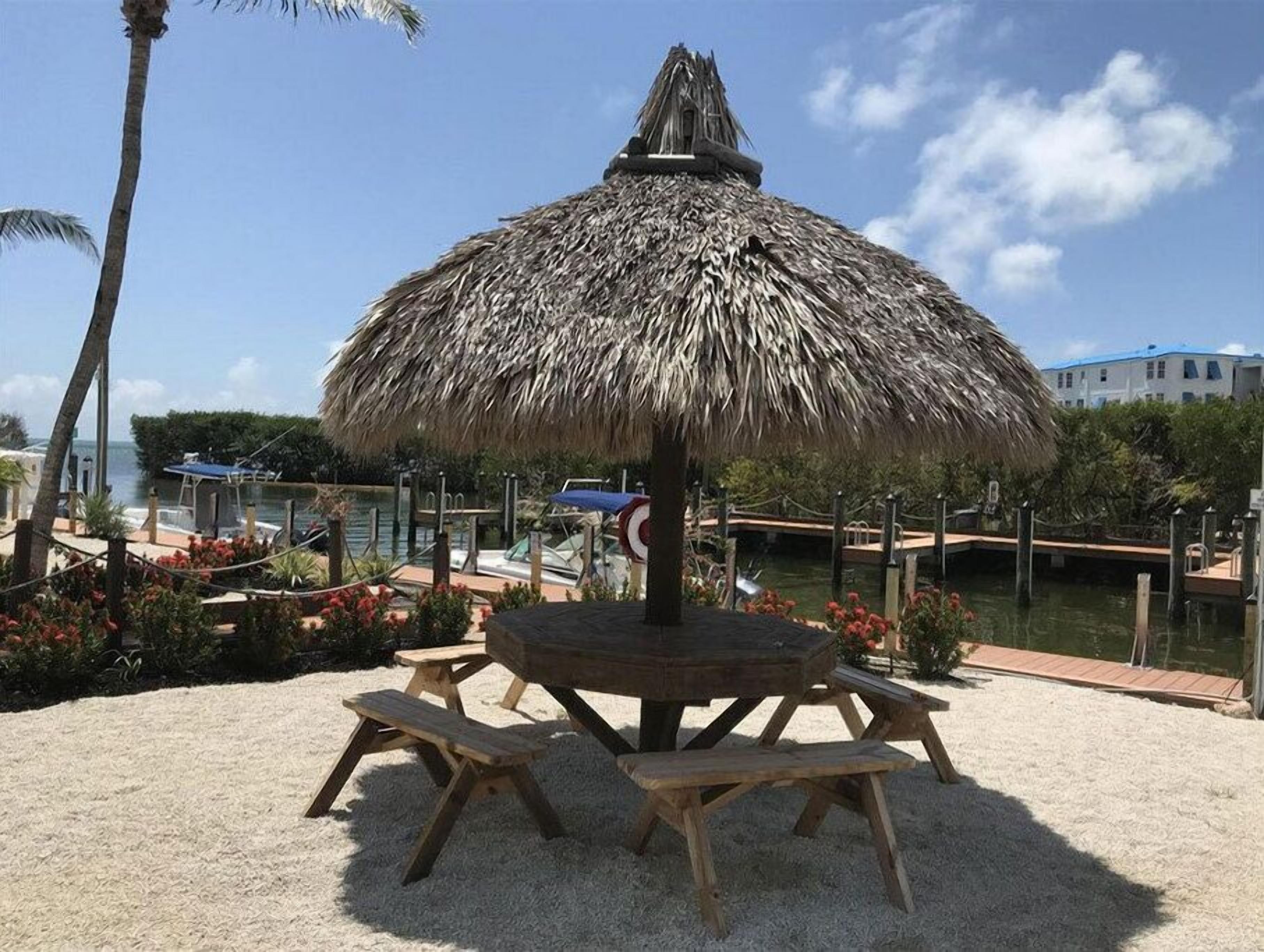 A thatched umbrella table with benches in a sandy area with a view of boats and water.