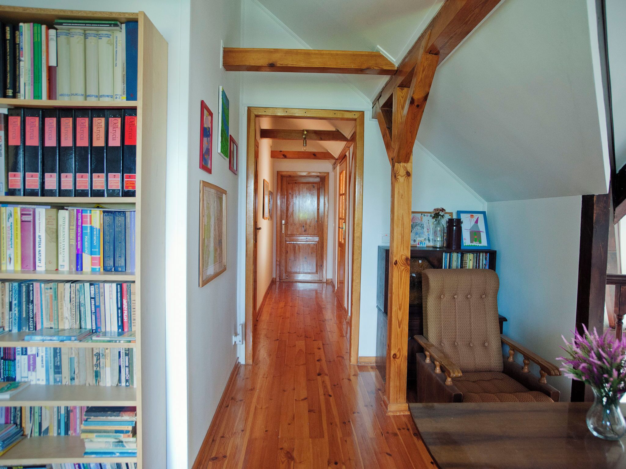 Een hal met houten boekenkasten links, een houten stoel rechts, en een houten trap die naar een andere kamer leidt.