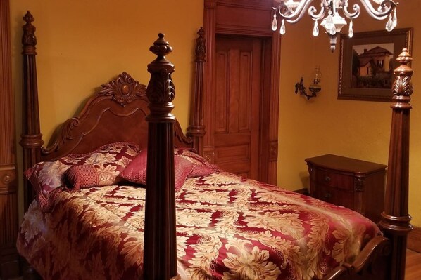 Una cama con dosel, un edredón floral, una mesita de noche de madera, una puerta de madera y un candelabro.