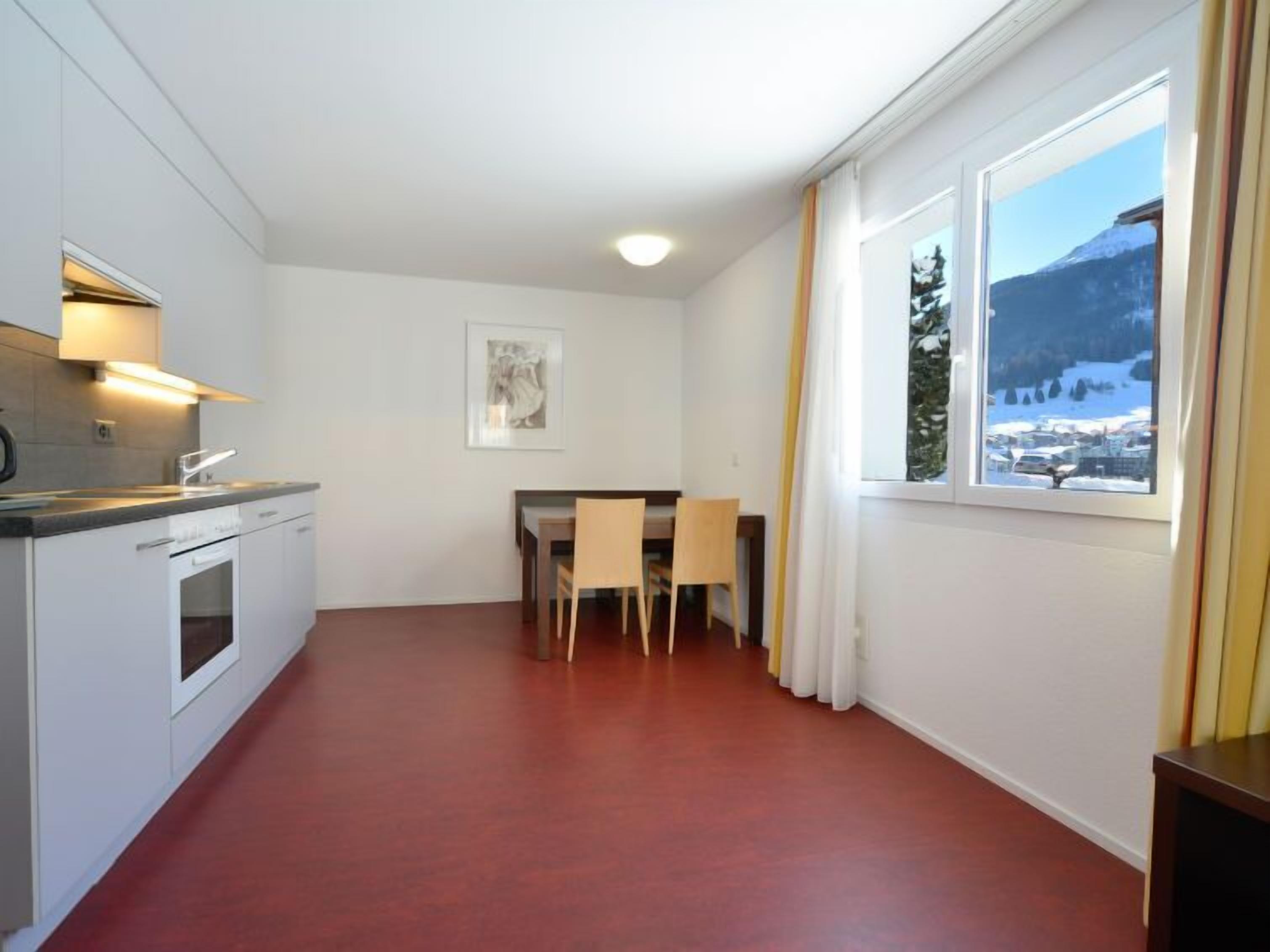 Une cuisine moderne avec des armoires blanches et un four encastré, un coin repas avec une table et des chaises, et une grande fenêtre offrant une vue sur les montagnes.