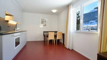 Une cuisine moderne avec des armoires blanches et un four encastré, un coin repas avec une table et des chaises, et une grande fenêtre offrant une vue sur les montagnes.