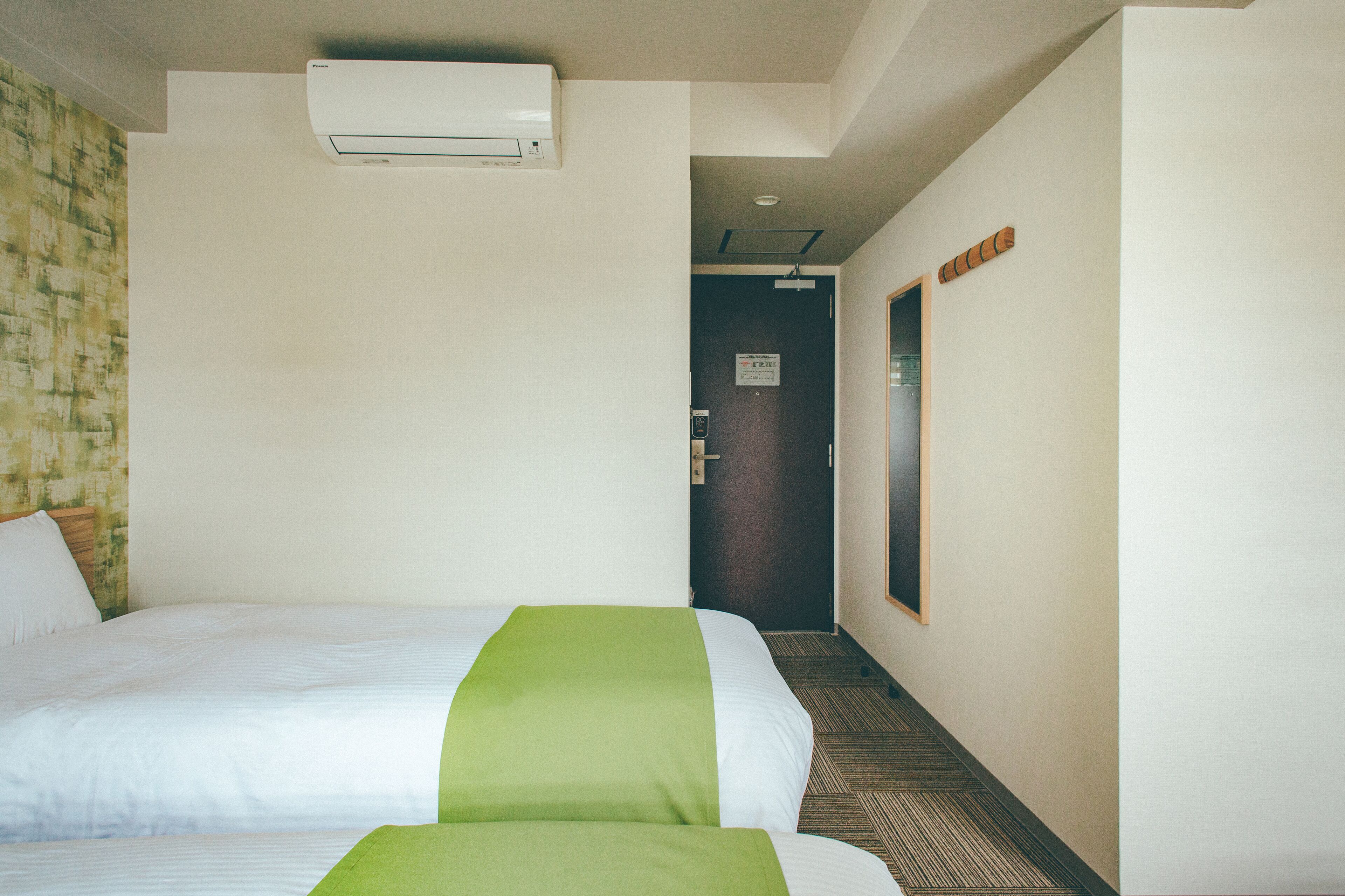 A hotel room with two beds, a green bedspread, a wall-mounted air conditioner, a door with a sign, and a carpeted floor.