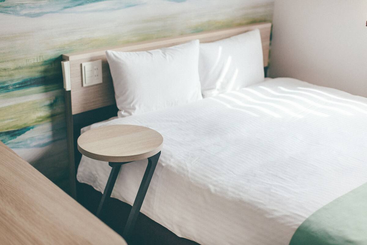 A neatly made bed with white linens, a round wooden nightstand, and a wall with a painting.