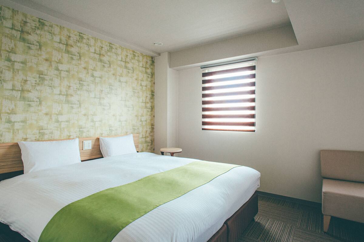 A hotel room with a large bed, a small table, a chair, and a window with blinds.