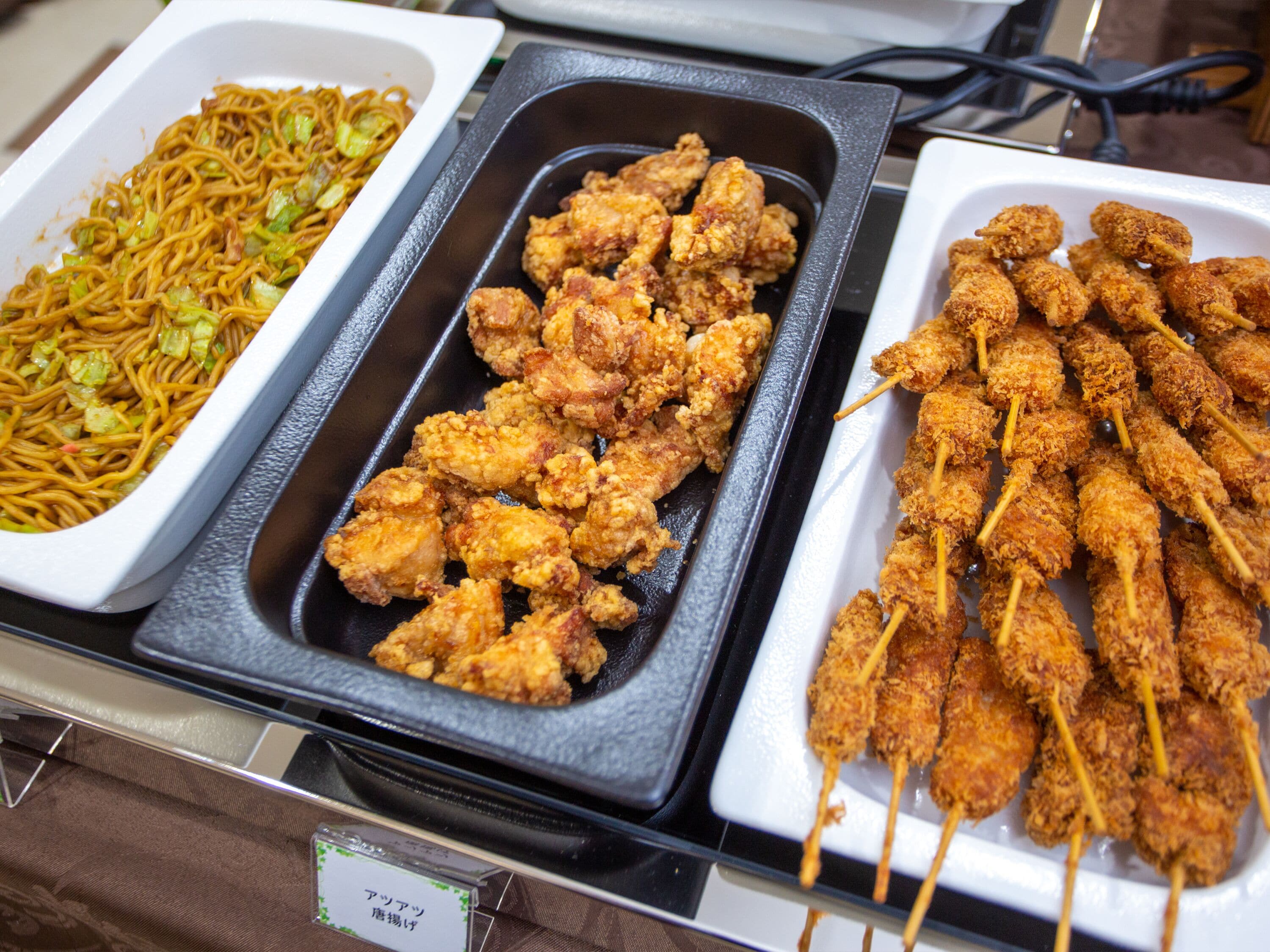 Un bufé con diversos platos, incluyendo fideos, pollo frito y brochetas.