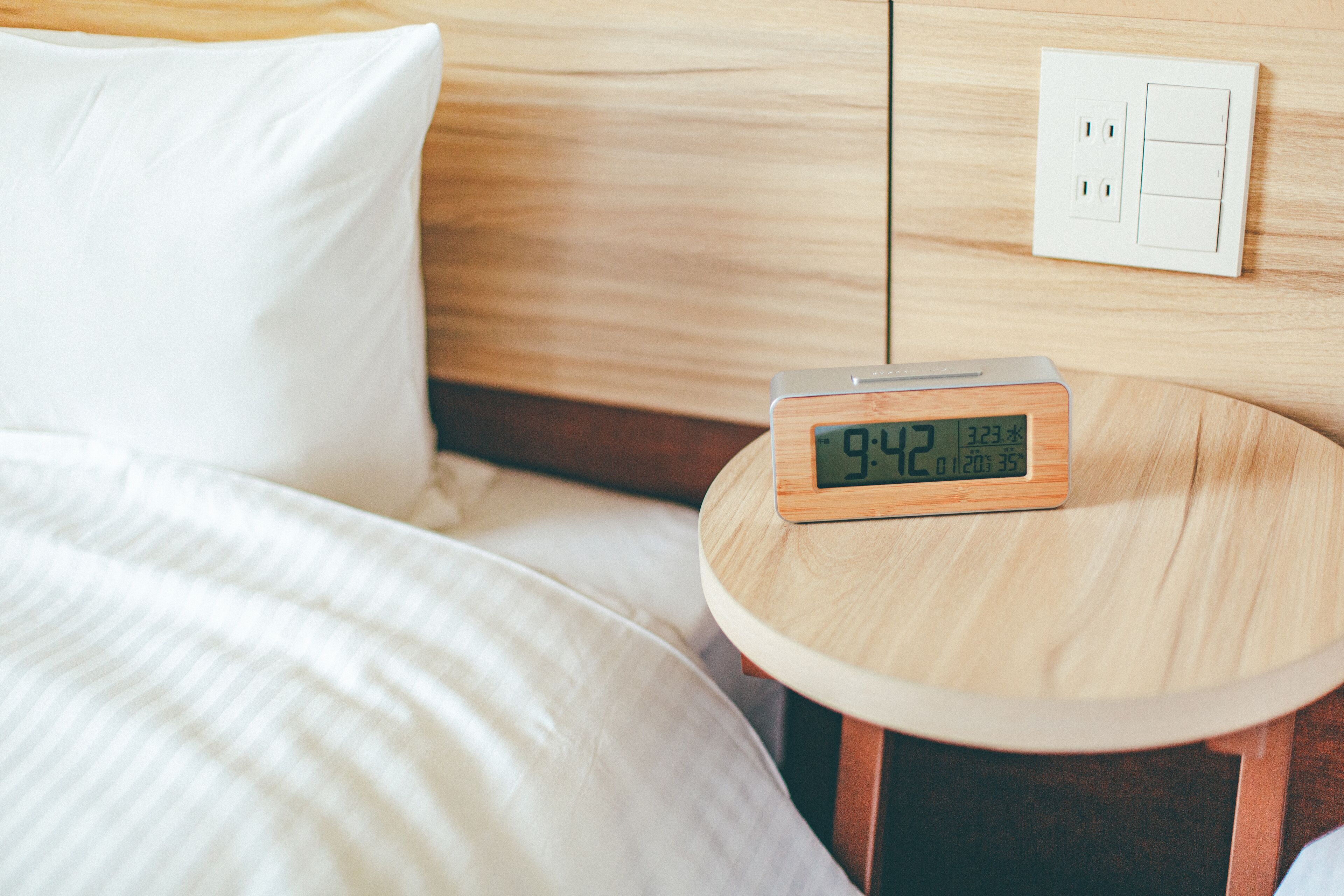 A bed with a white pillow and a round wooden nightstand with a digital clock displaying the time.