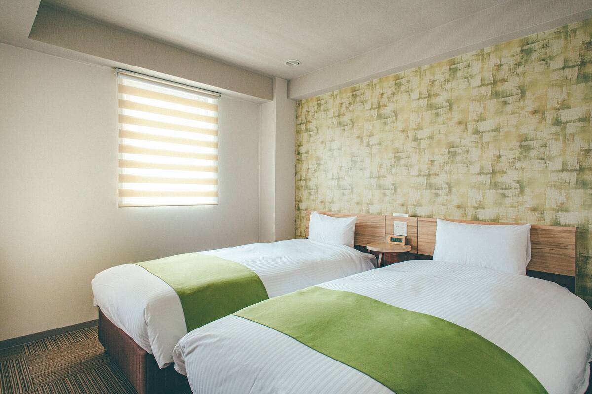 A hotel room with two single beds, a window with blinds, a headboard with a wooden panel, and a patterned wallpaper on the wall.