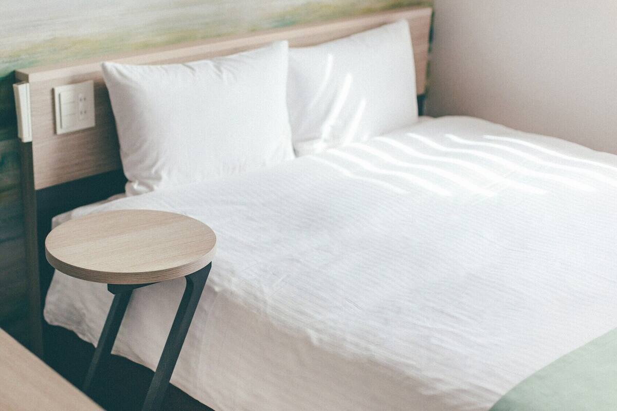 A neatly made bed with white bedding, a round wooden side table, and a headboard with a light switch.