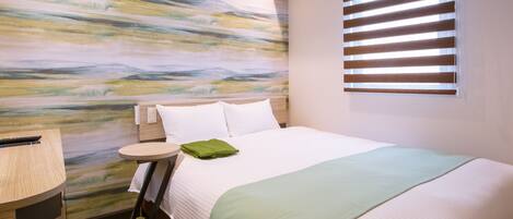 Una habitación de hotel con cama, mesita de noche, lámpara y ventana con persianas.