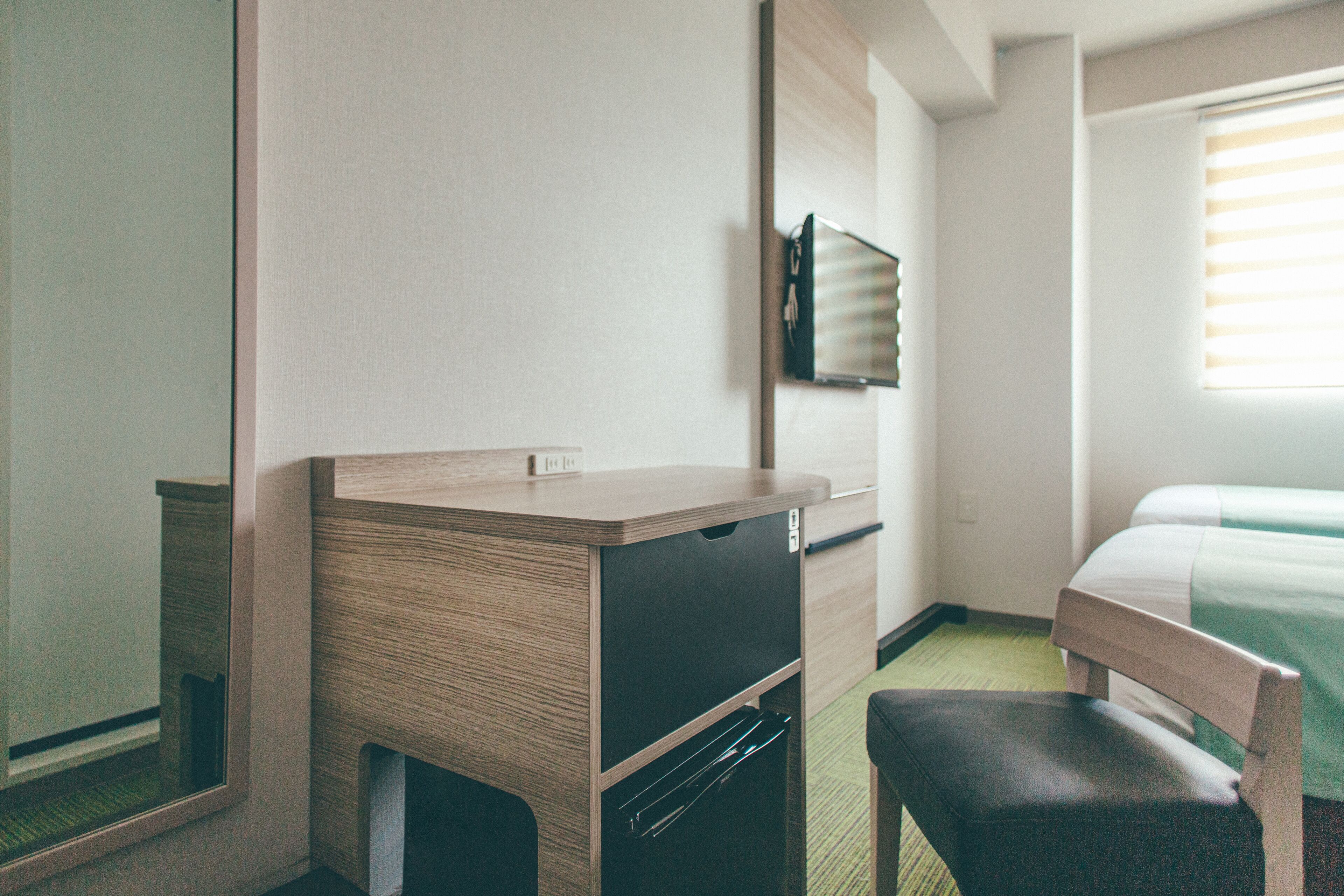 A compact hotel room with a wooden desk, a chair, a bed with a green bedspread, and a mirror on the wall.