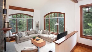 A modern living room with a sectional sofa, a coffee table, and large windows offering a view of the forest. - Ski-access retreat w/ hot tub & fireplace (Whistler)