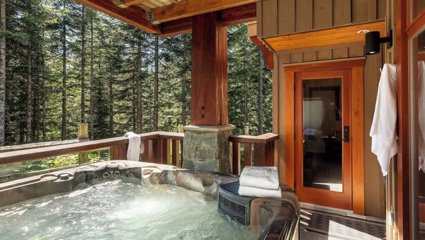 A hot tub on a wooden deck with a forest view, a stone pillar, and a door with a towel hanging on it. - Ski-access retreat w/ hot tub & fireplace (Whistler)