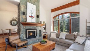 A modern living room with a stone fireplace, a large window offering a view of trees, and a cozy seating area with a sofa and chairs. - Ski-access retreat w/ hot tub & fireplace (Whistler)