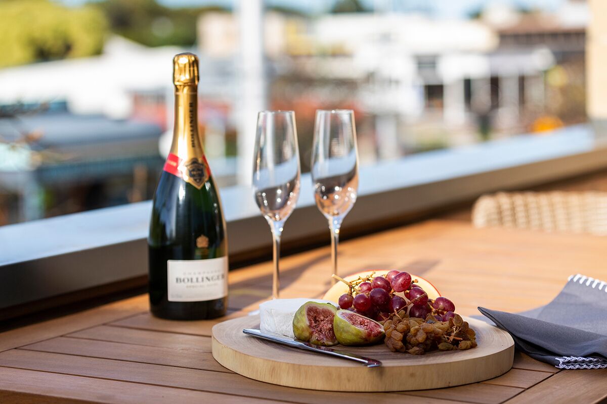 A bottle of Bollinger wine, two wine glasses, and a wooden board with cheese, grapes, and nuts on a terrace with a view.