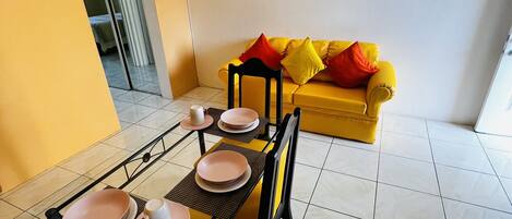 A dining area with a table set for four, chairs, and a yellow sofa with red cushions in the background.