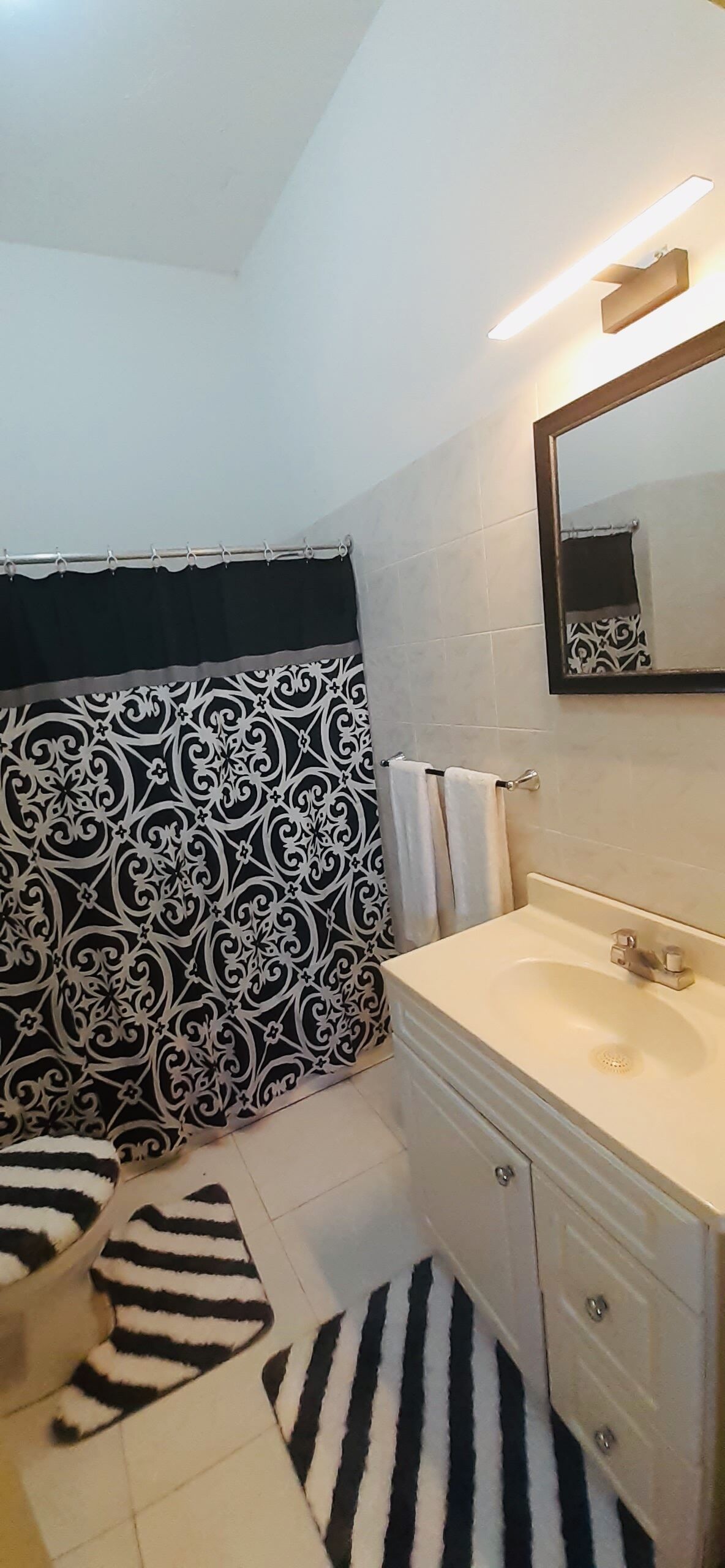 Un baño con lavamanos blanco, un espejo, toallero y cortina de ducha con estampado blanco y negro.