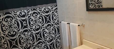 Un baño con lavamanos blanco, un espejo, toallero y cortina de ducha con estampado blanco y negro.
