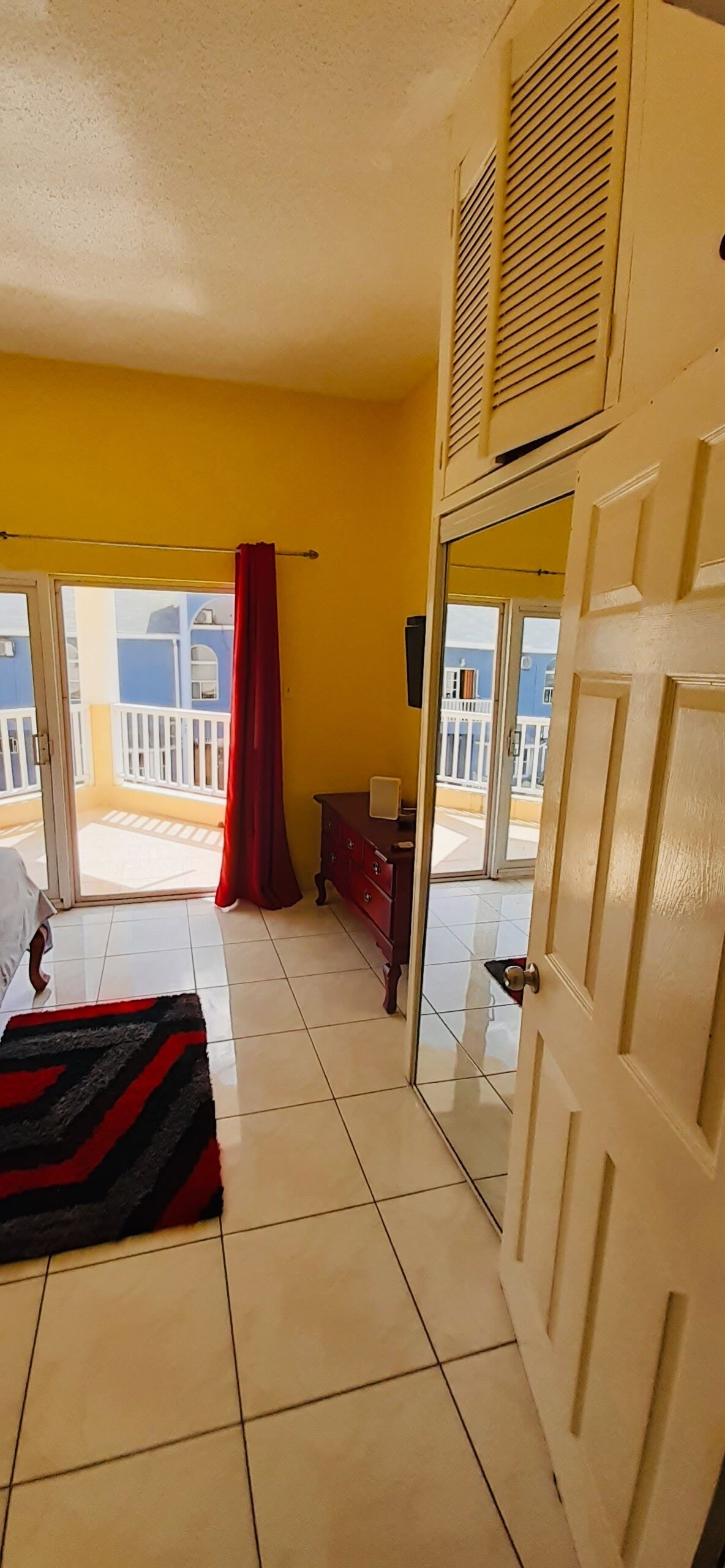 Una camera d'albergo con un letto, un tappeto a righe rosse e nere, un comò in legno e una porta scorrevole in vetro che dà su un balcone con vista sugli edifici.