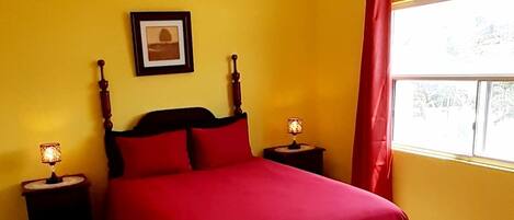 A bedroom with a red bed, two bedside tables with lamps, a framed picture on the wall, and a window with curtains.