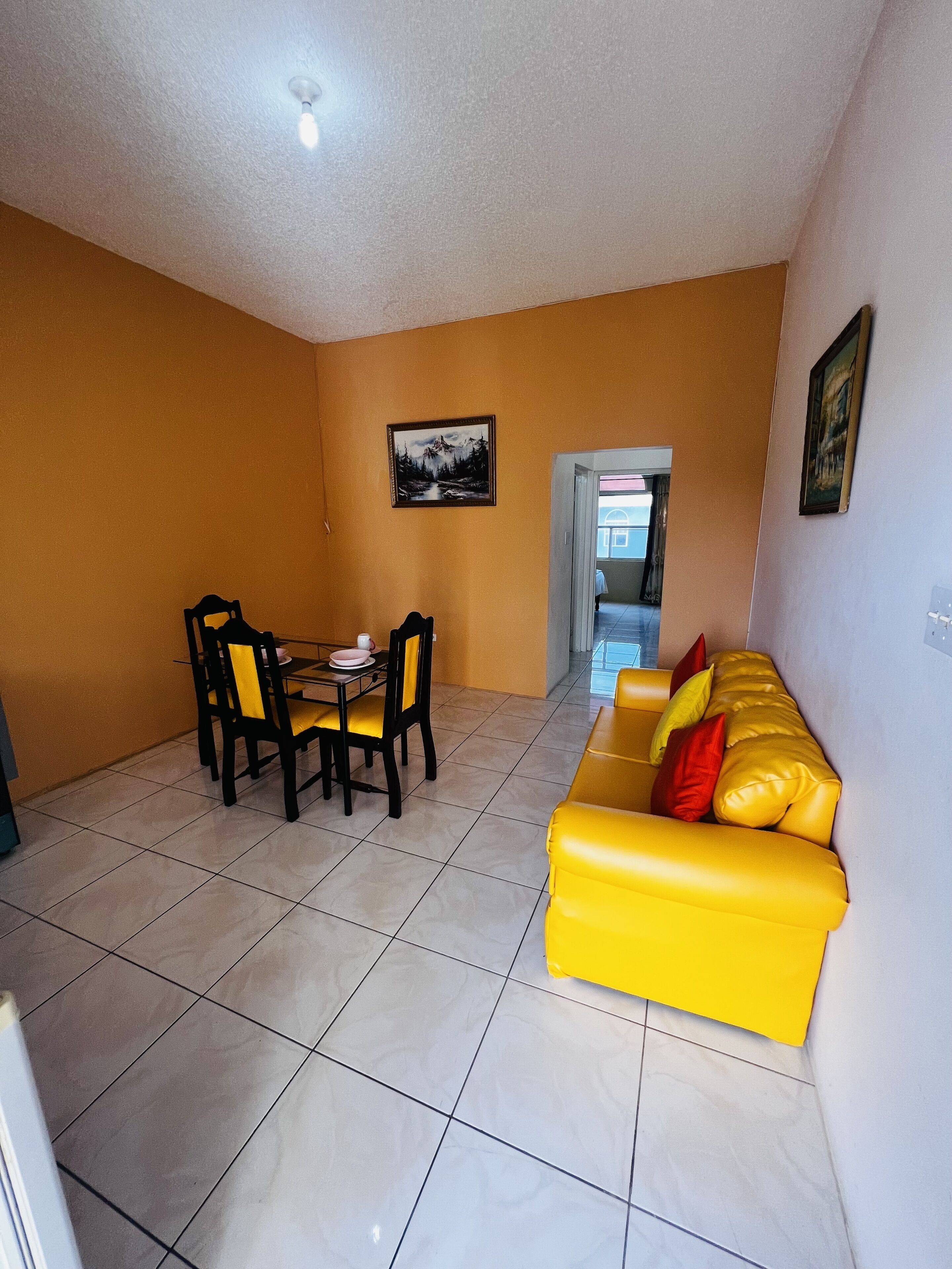 A dining area with a table and chairs, a yellow sofa with pillows, and a framed picture on the wall.