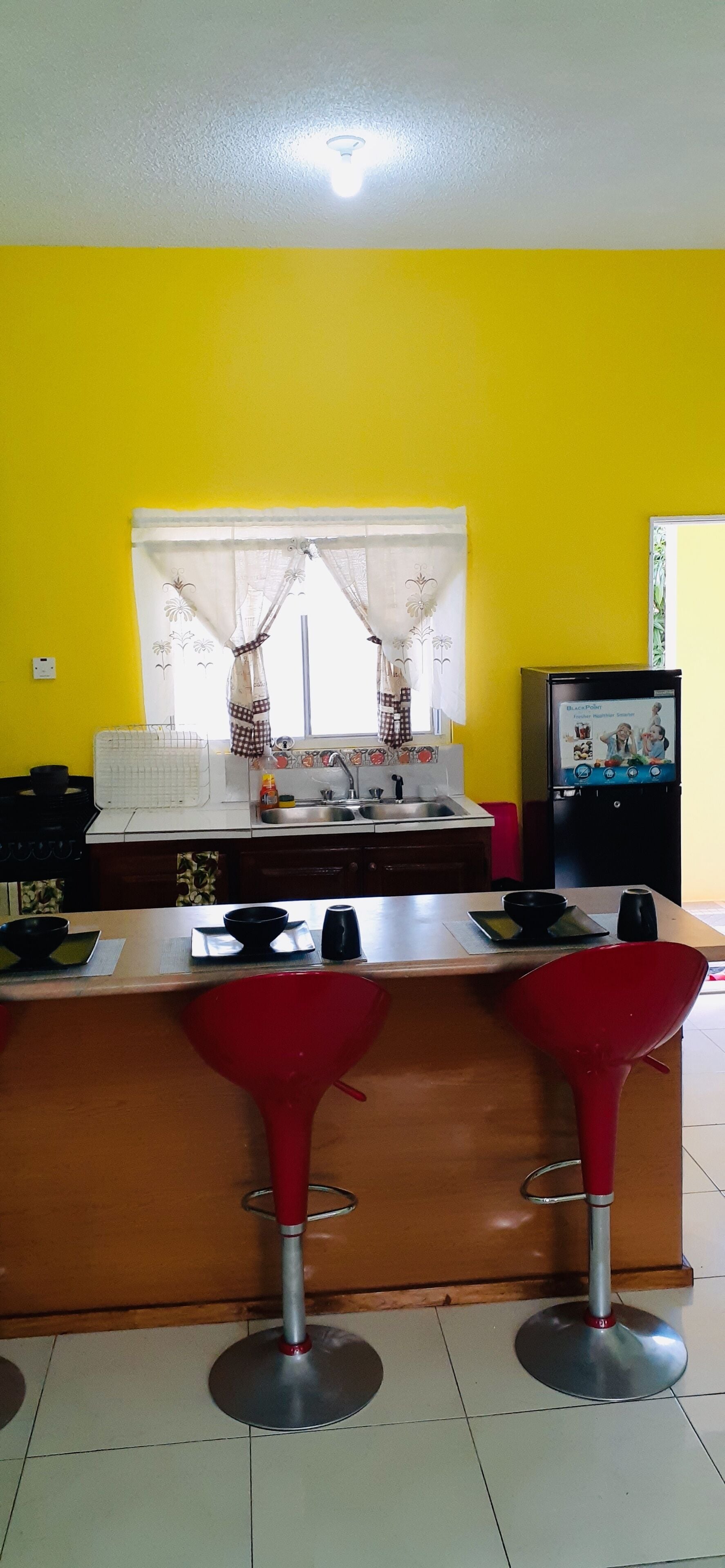 Una cocina con pared amarilla, ventana con cortinas, un fregadero, refrigerador y una barra con taburetes.