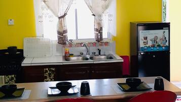 Una cocina con pared amarilla, ventana con cortinas, un fregadero, refrigerador y una barra con taburetes.