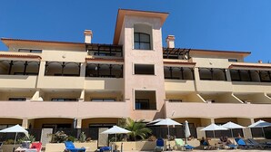 Un hôtel à plusieurs étages doté de balcons, d’un espace piscine avec des chaises longues et des parasols, et un ciel d’un bleu limpide.