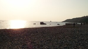 Une plage de galets, le soleil se couchant sur l’eau, un paysage rocheux et quelques personnes qui se promènent.