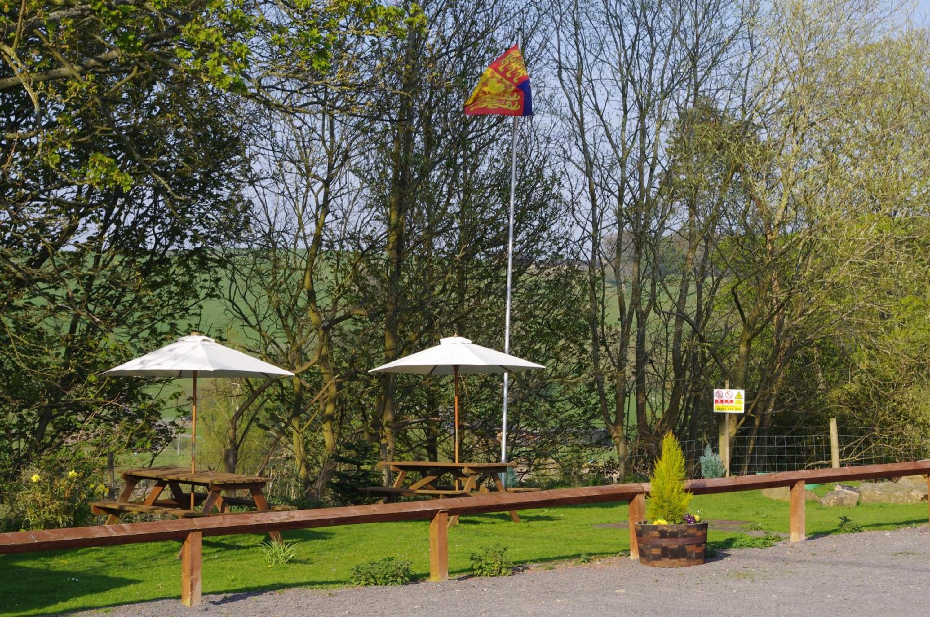 A picnic area with two wooden tables and white umbrellas, surrounded by trees and a flag.