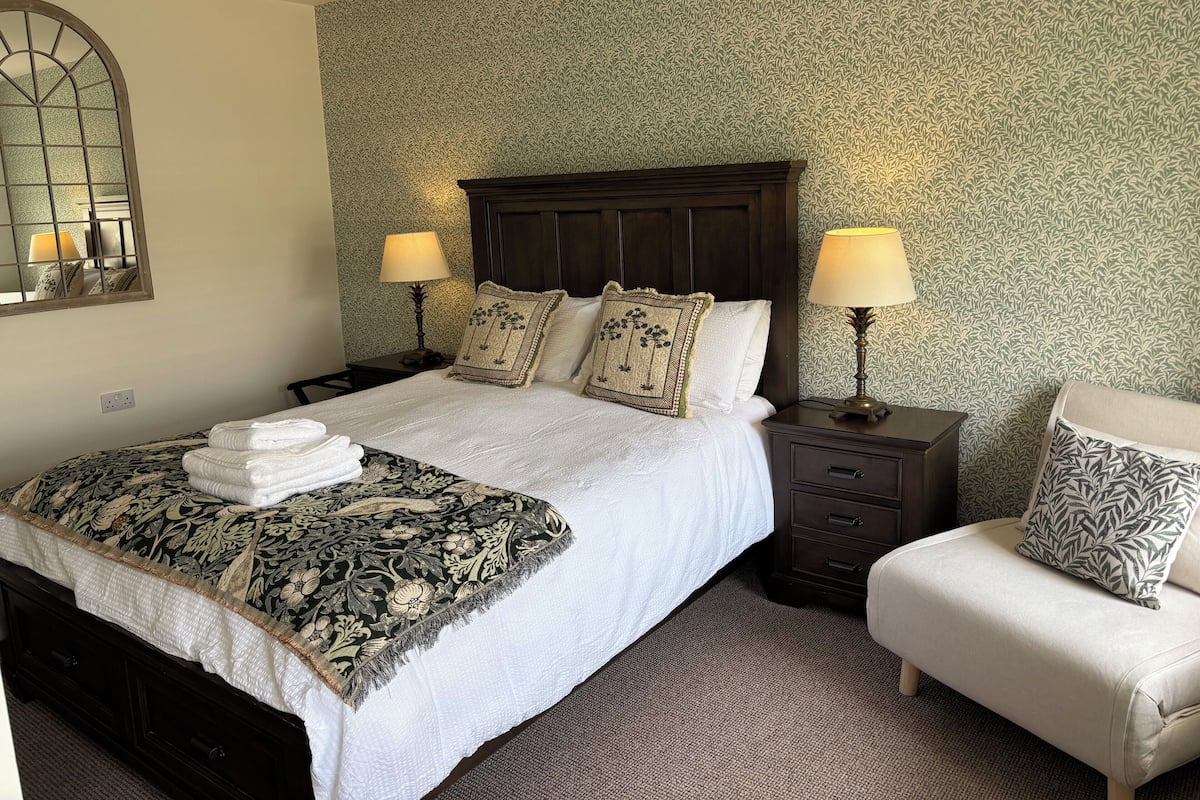 Un lit bien fait, recouvert d’une couvre-lit à motifs floraux, deux lampes de chevet, une table de chevet, une chaise et un mur décoré d’un papier peint à motifs.