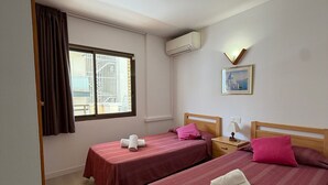 Habitación de hotel con dos camas, una ventana con cortinas, un equipo de aire acondicionado, una mesita de noche, una lámpara y un cuadro en la pared.